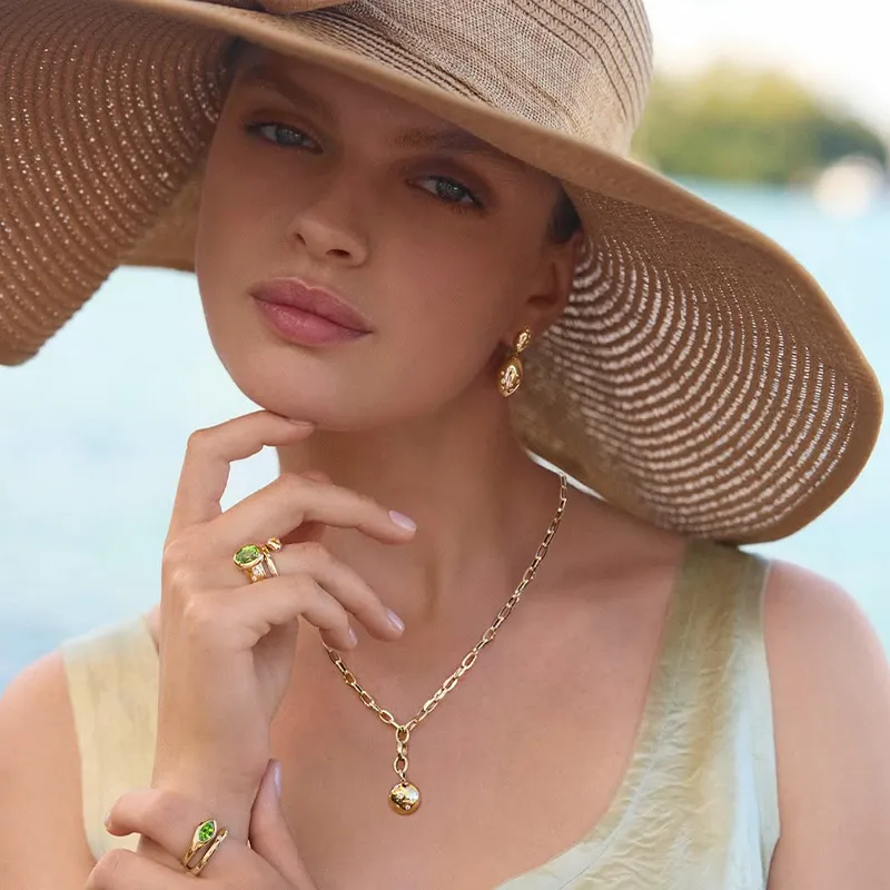 A woman wearing a wide-brimmed hat, gold jewelry, and a light dress by the water.