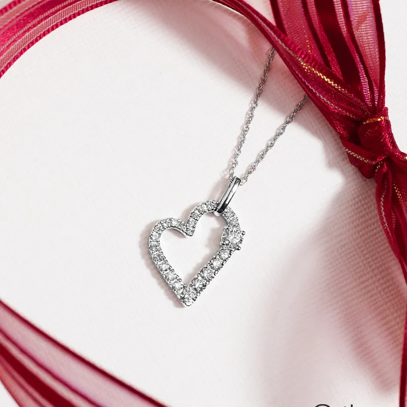 Silver heart pendant with diamonds on a delicate chain, displayed with a red ribbon backdrop.