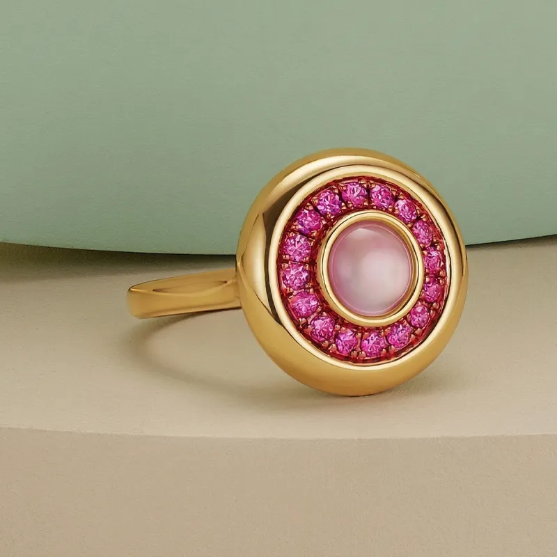 A gold ring with a pink pearl center, surrounded by pink gemstones, on a neutral background.