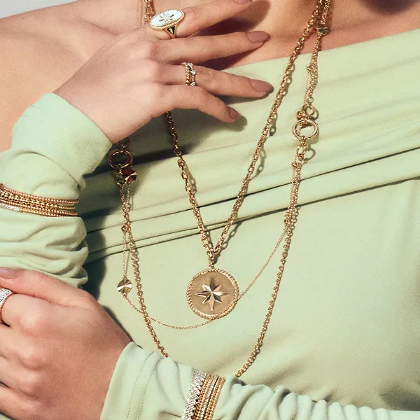 Woman in green dress wearing layered gold necklaces and rings. Click to explore more necklaces at R. Gregory Jewelers.