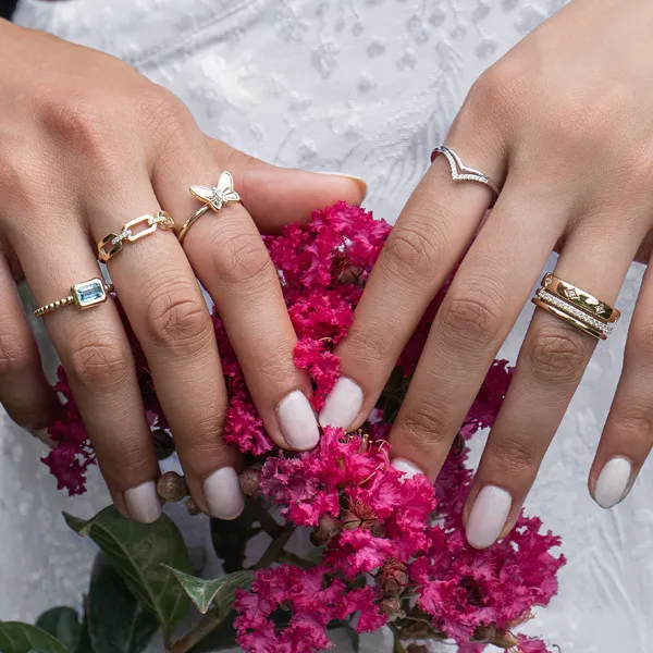 Hands with various gold rings holding pink flowers. Click to explore fashion rings at R. Gregory Jewelers.
