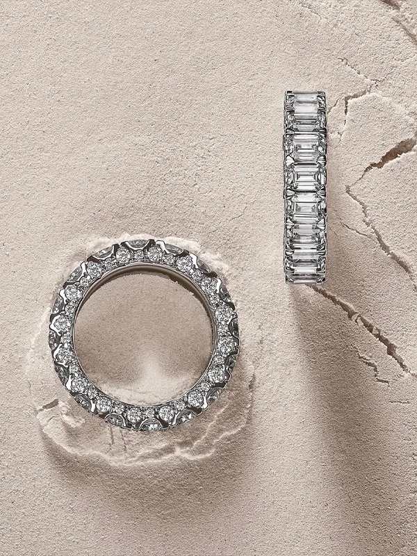 Two elegant wedding bands, one with round diamonds and the other with rectangular stones, displayed on a textured surface.