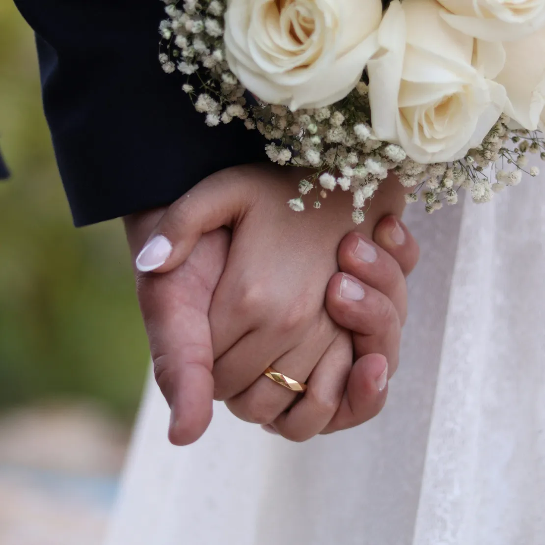 Hands holding with wedding bands, bouquet of white roses nearby.