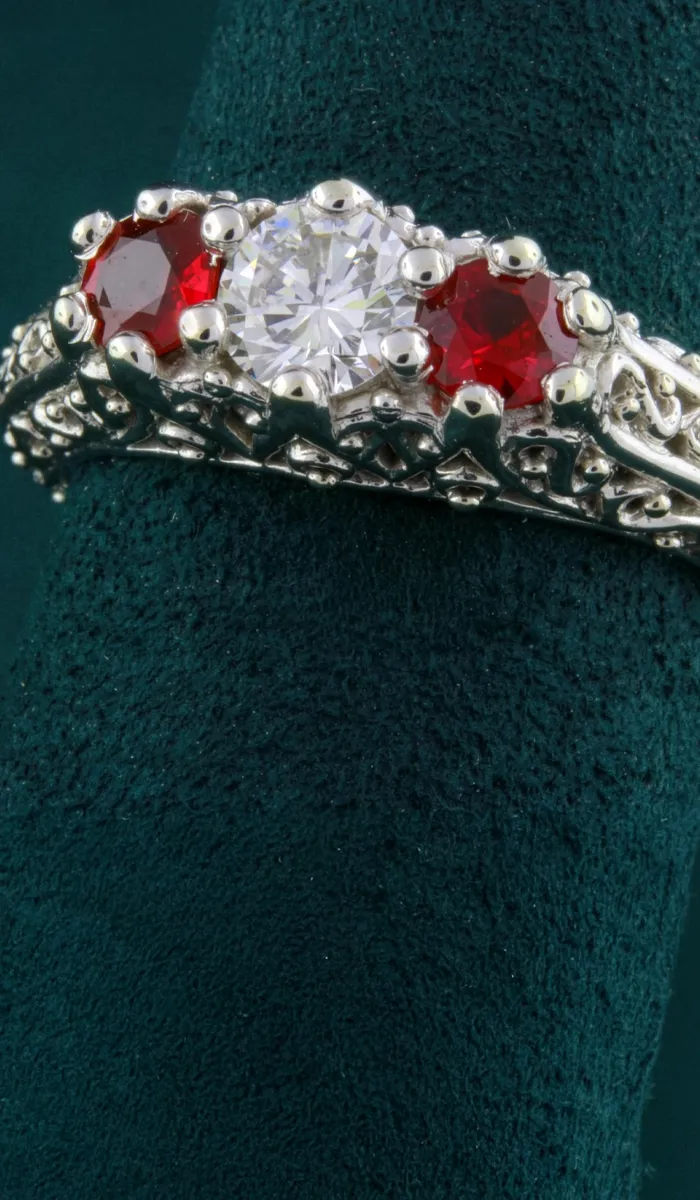 Elegant silver ring featuring a central diamond flanked by two red gemstones, displayed on a green velvet stand.