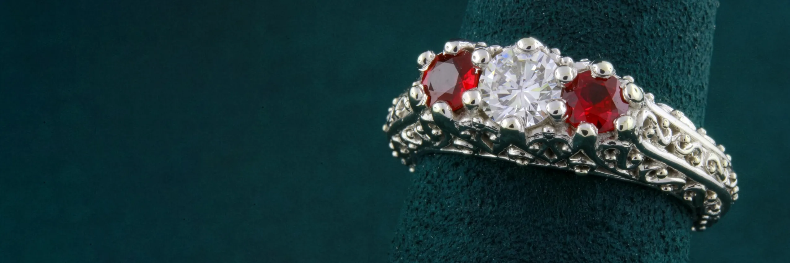 Elegant silver ring featuring a central diamond flanked by two red gemstones, displayed on a green velvet stand.