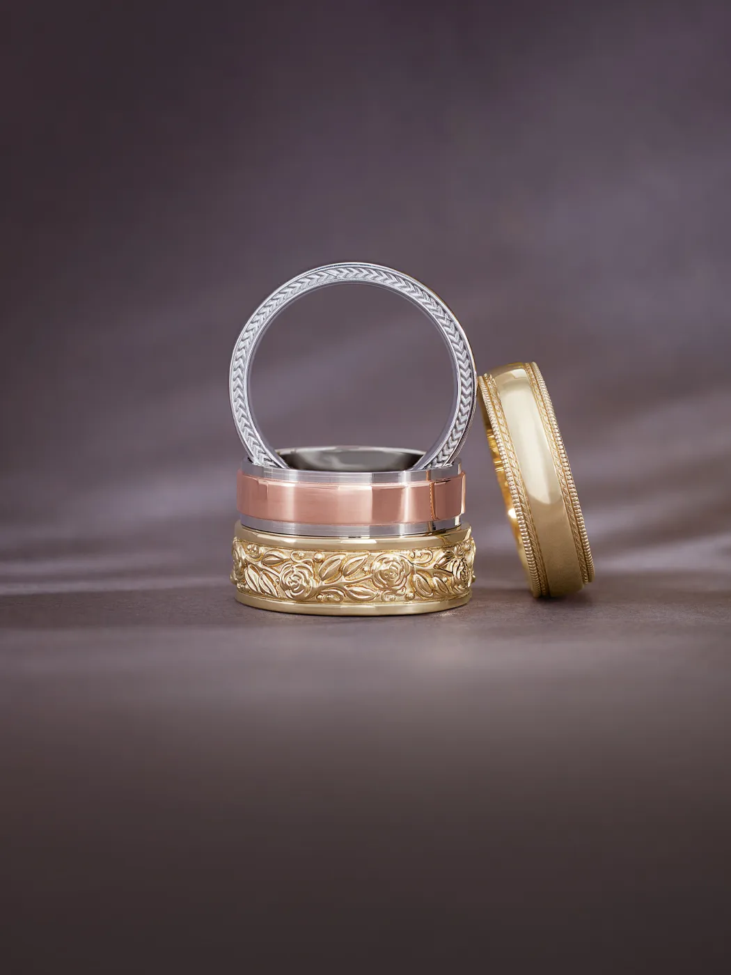 Three elegant men's wedding bands stacked: silver, rose gold, and gold with floral engraving.