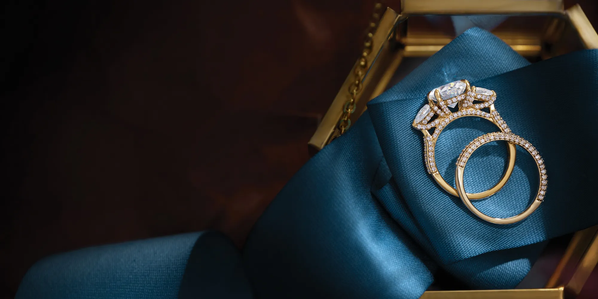 Elegant gold engagement and wedding rings on a teal ribbon, inviting you to shop bridal sets.
