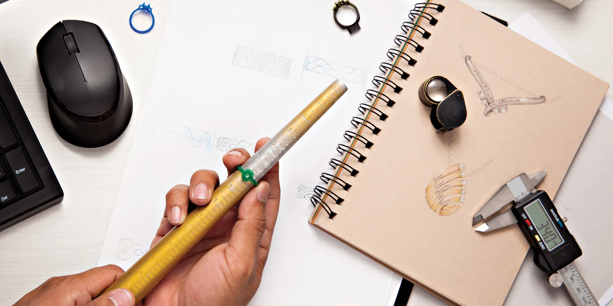 A hand holding a measuring tool over sketches and jewelry design materials on a workspace.