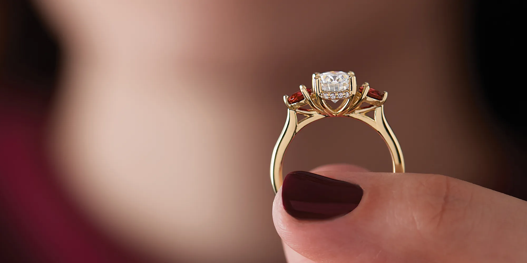 A woman holds a stunning gold engagement ring with a central diamond and red gemstones.