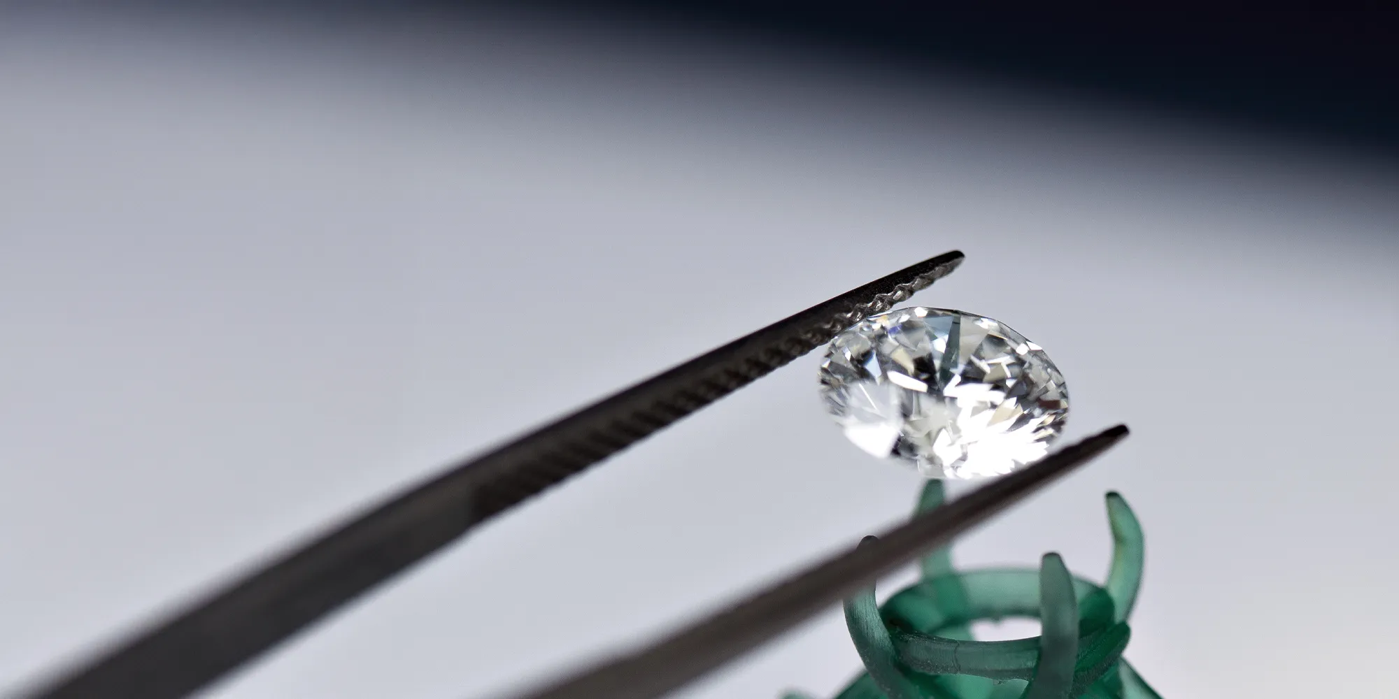 A diamond held by tweezers above a green ring mount, showcasing its brilliance and clarity.