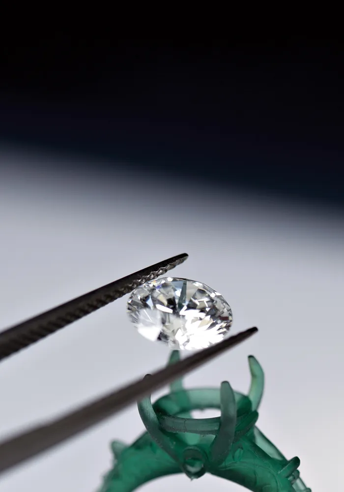 A diamond held by tweezers above a green ring mount, showcasing its brilliance and clarity.