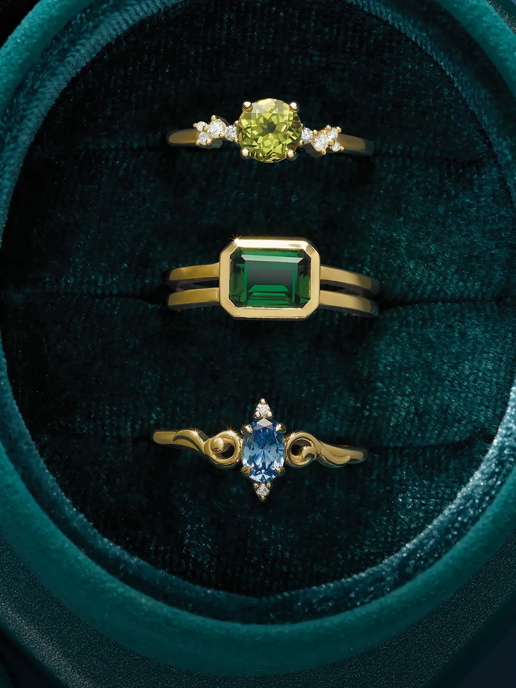 Three elegant rings featuring a yellow gemstone, a green emerald cut stone, and a blue oval stone, displayed in a velvet box.