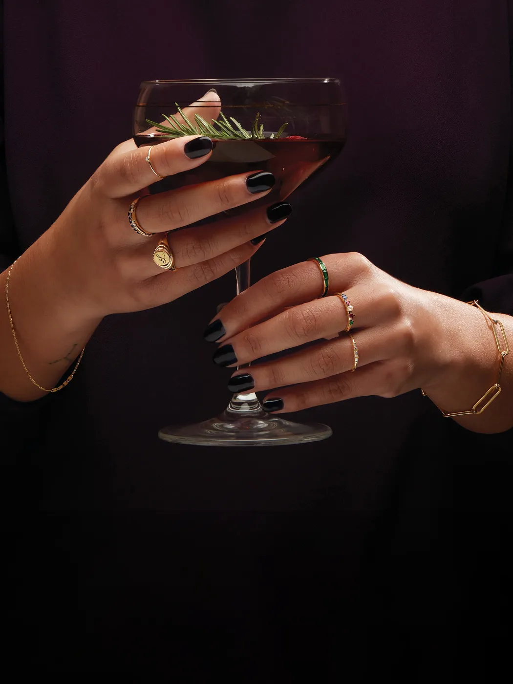 Elegant hands holding a cocktail, adorned with various gold rings and black nails.