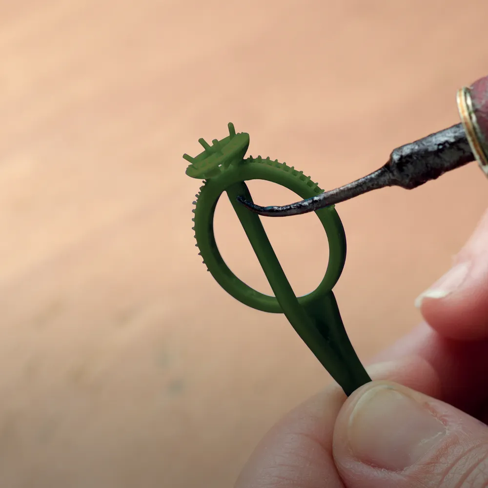 A person uses a tool to shape a green wax ring model for jewelry making.
