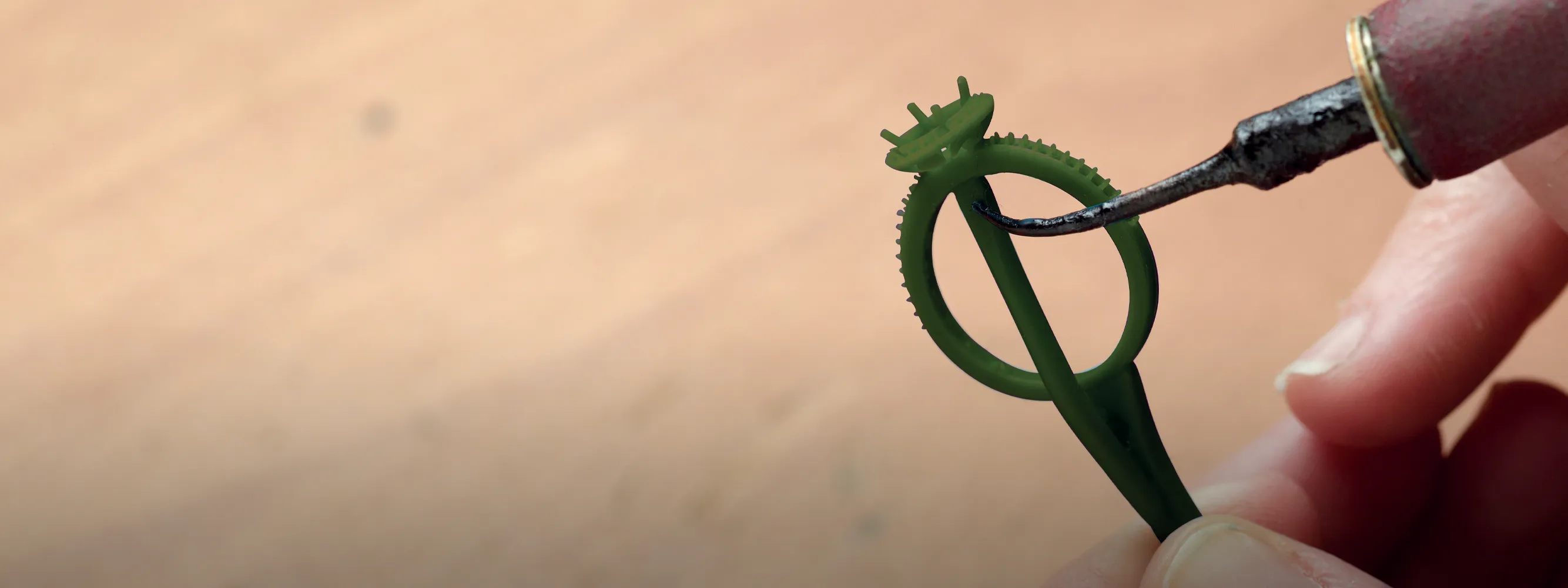 A person uses a tool to shape a green wax ring model for jewelry making.