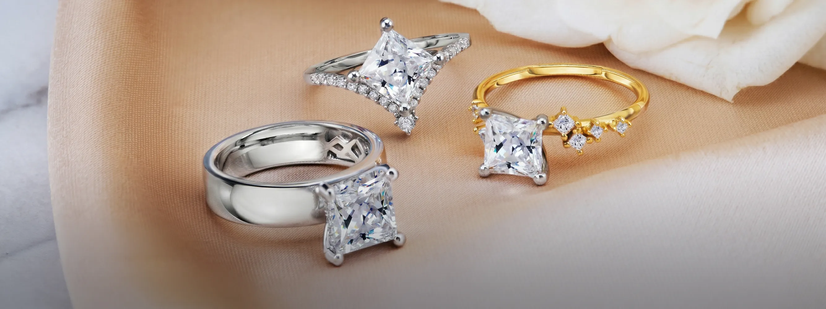 Three elegant engagement rings with large square diamonds on a soft fabric background.