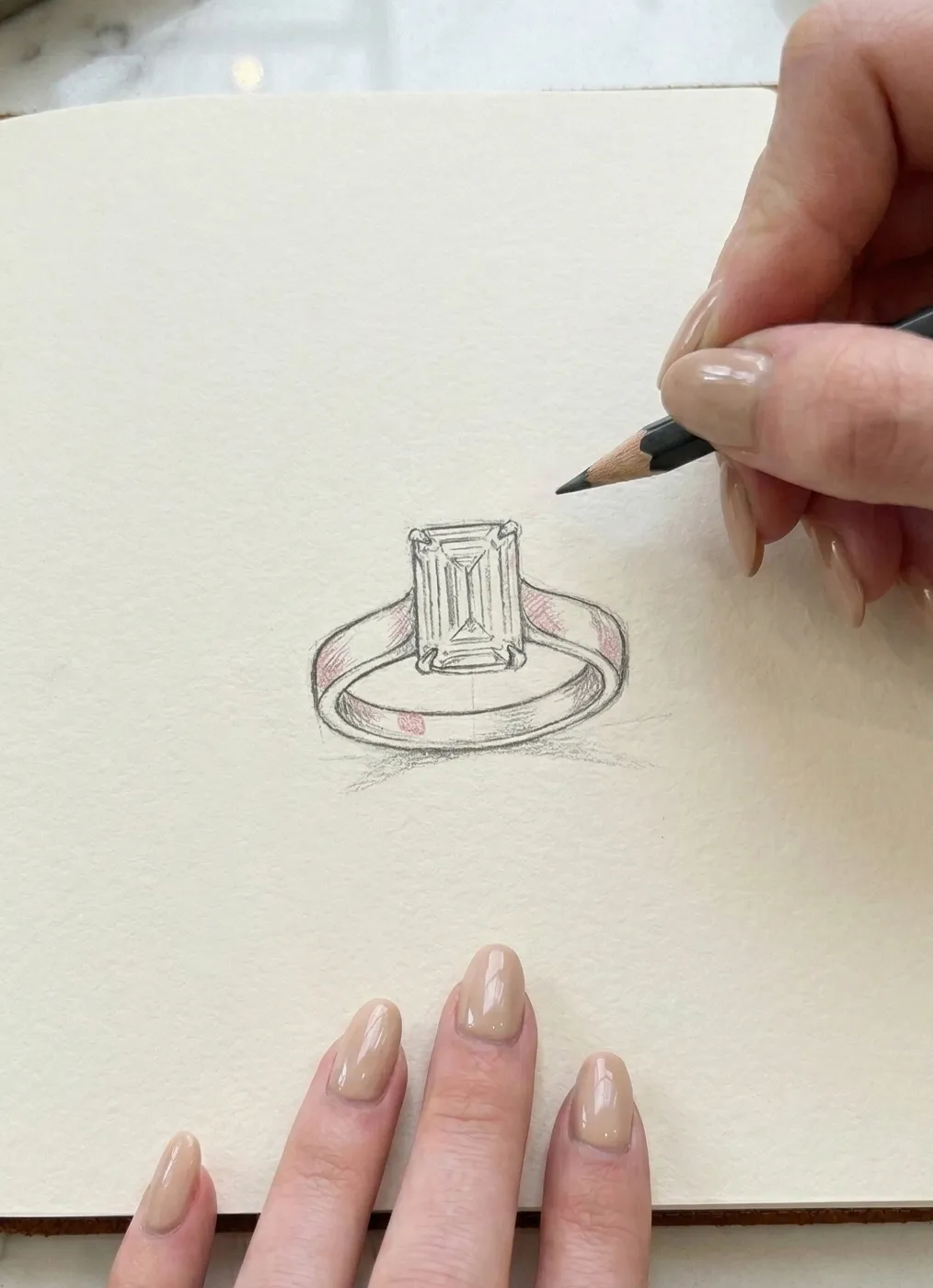 A hand sketches an emerald-cut diamond ring on paper with a pencil.