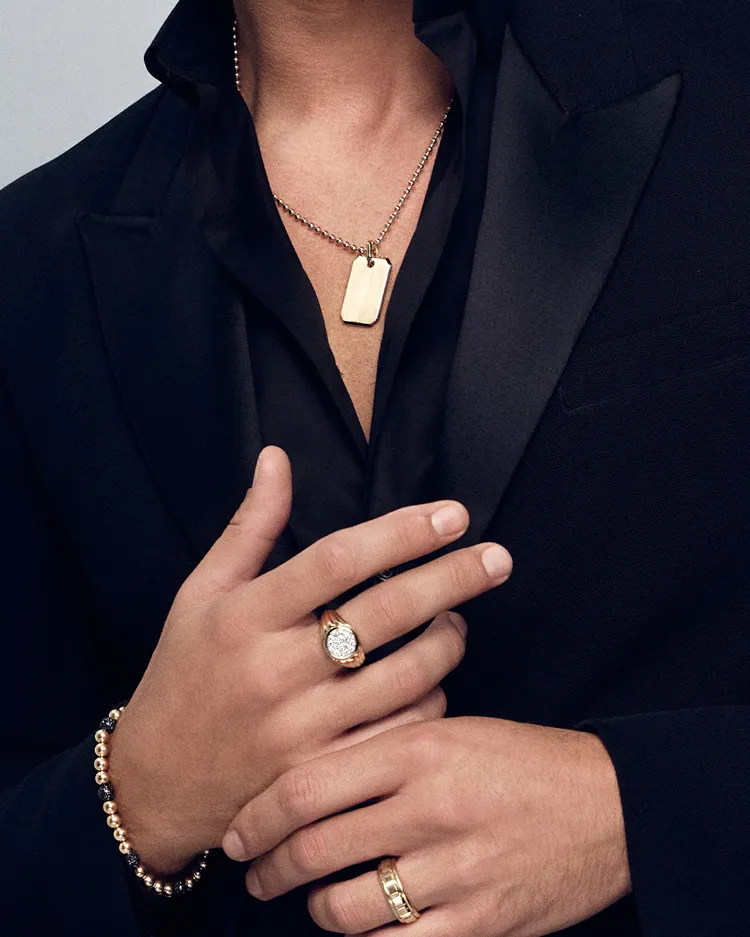 Close-up of a man in a black suit showcasing gold jewelry, including a dog tag necklace and multiple rings.