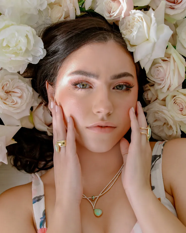 A woman with jewelry poses among white roses, showcasing Sather Jewelry's elegant designs.