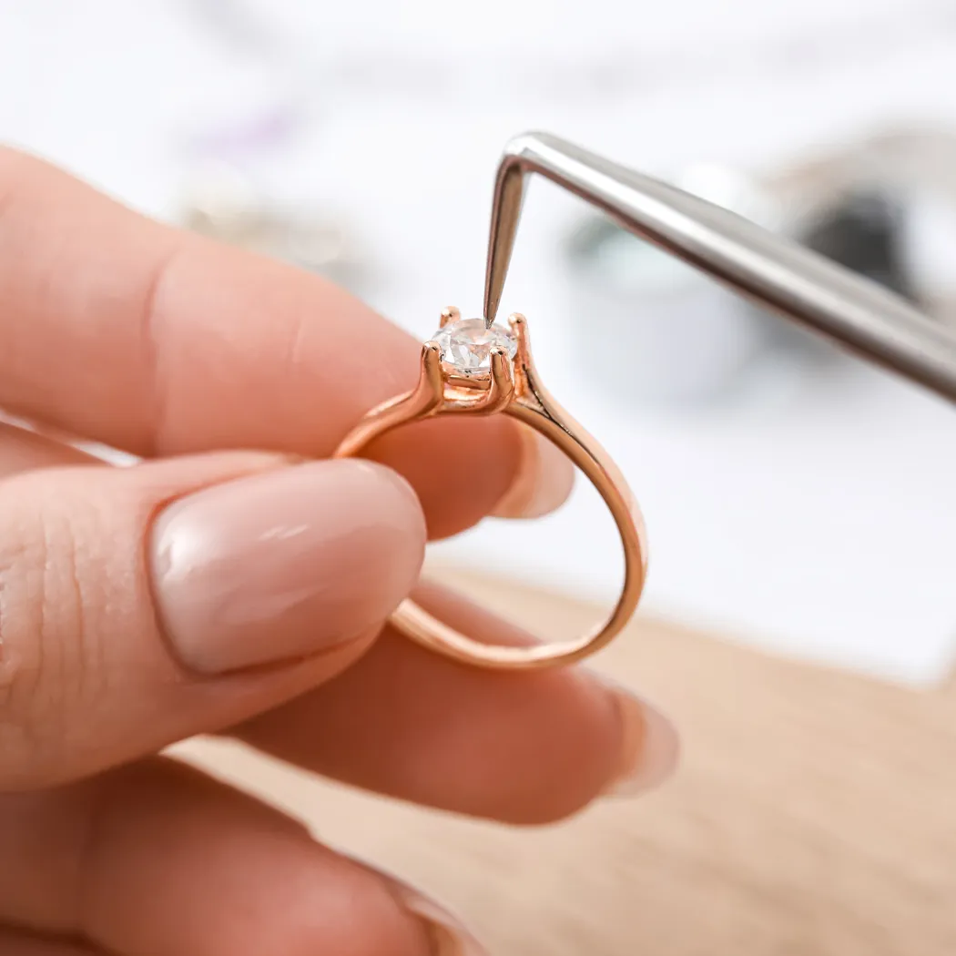Hand holding a rose gold engagement ring with a diamond, being adjusted with a tool.