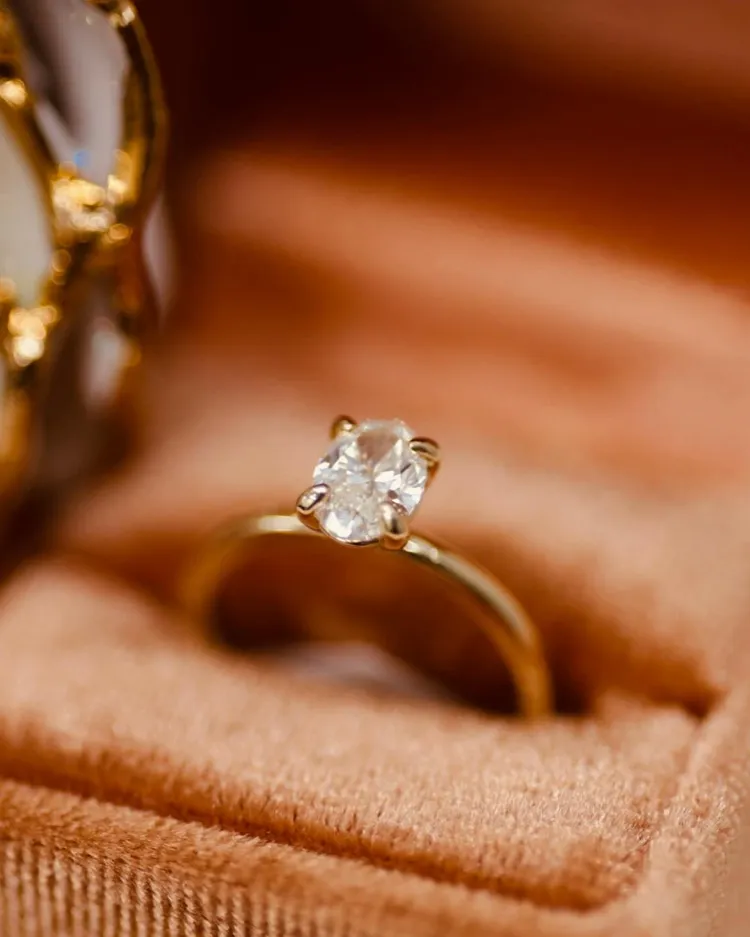 Elegant gold engagement ring with a sparkling oval diamond, displayed on a soft, plush background.