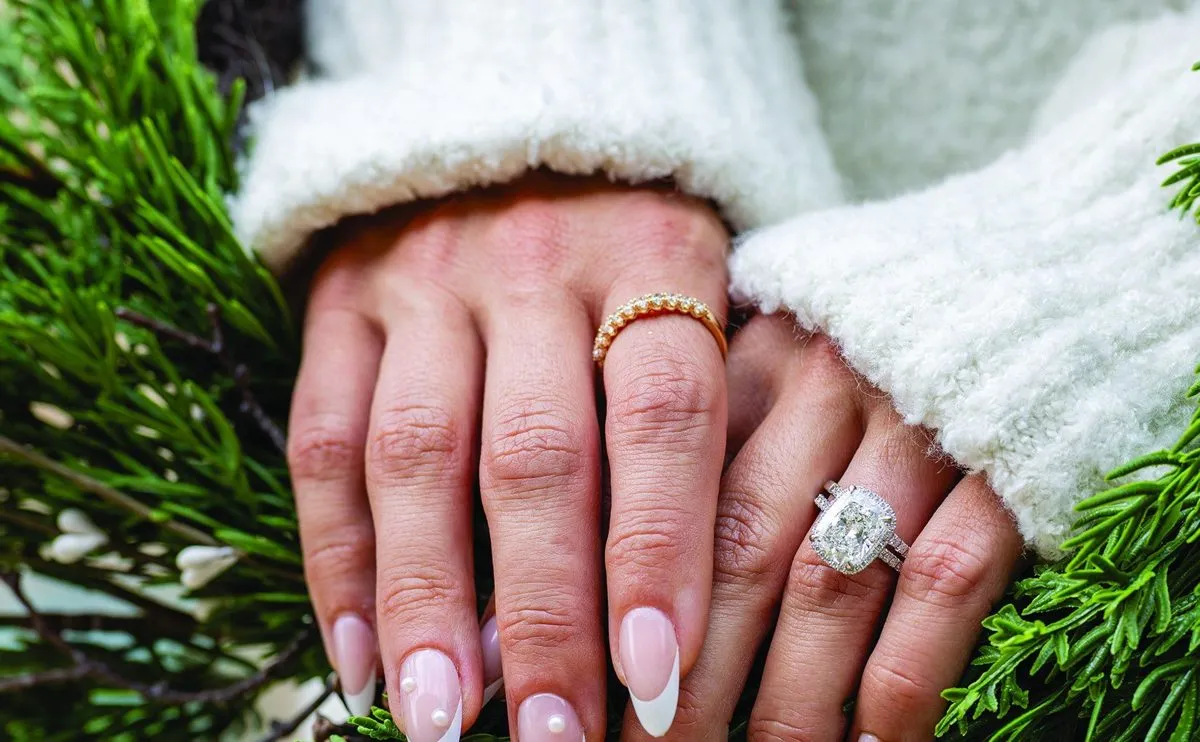 Hands in a cozy white sweater wearing a cushion-cut diamond halo ring and a gold band, resting on evergreen