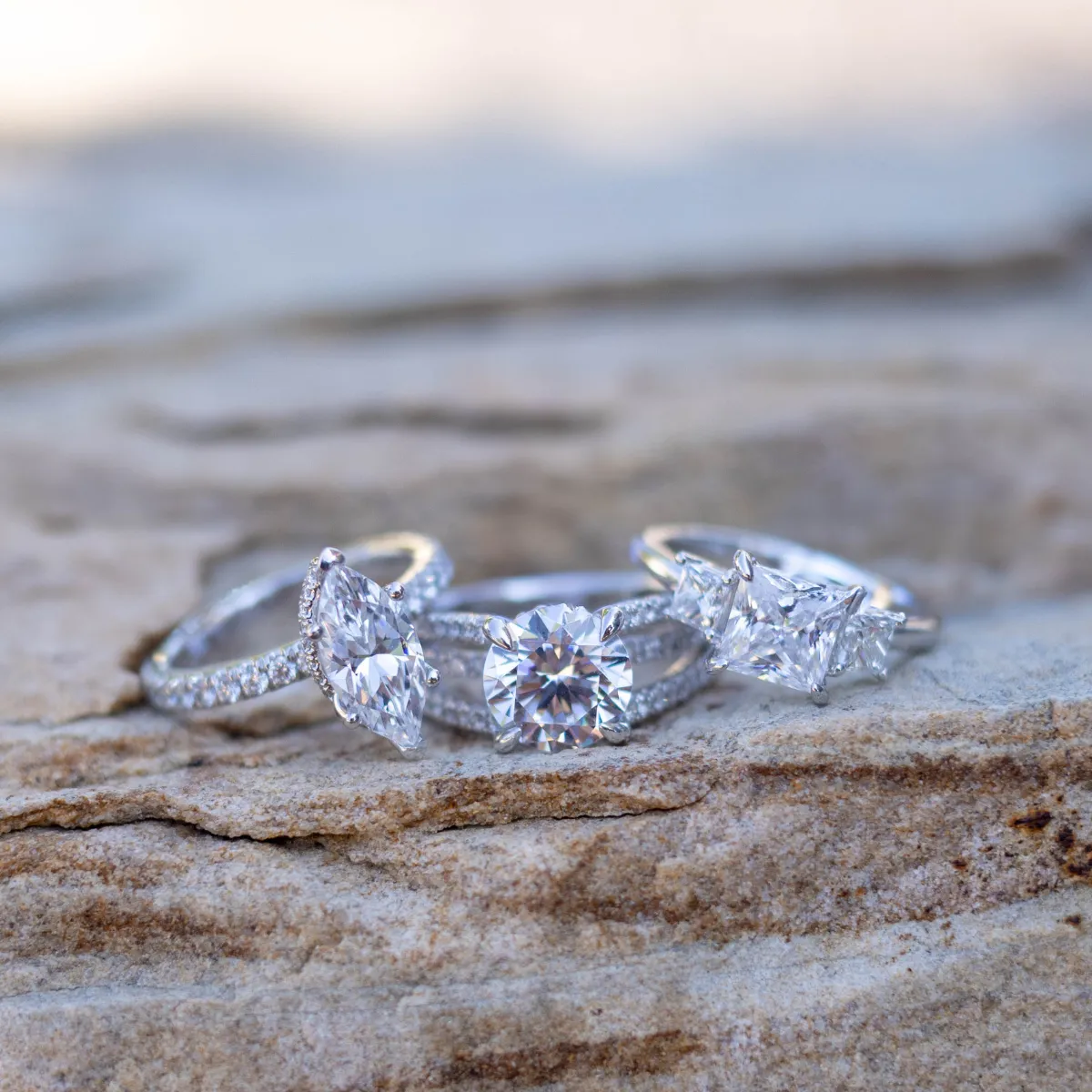 Three elegant engagement rings with various diamond cuts displayed on a stone surface. Visit Sather's Leading Jewelers.