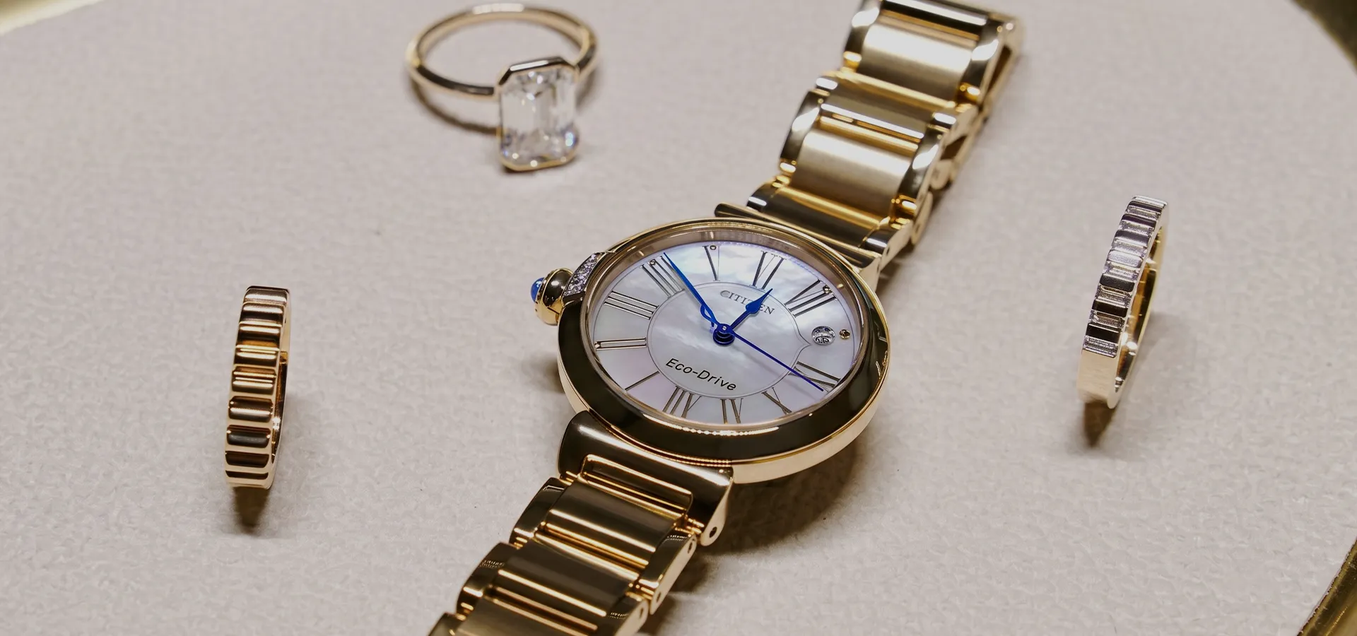 Gold watch with blue hands and Eco-Drive text, surrounded by rings on a textured surface.