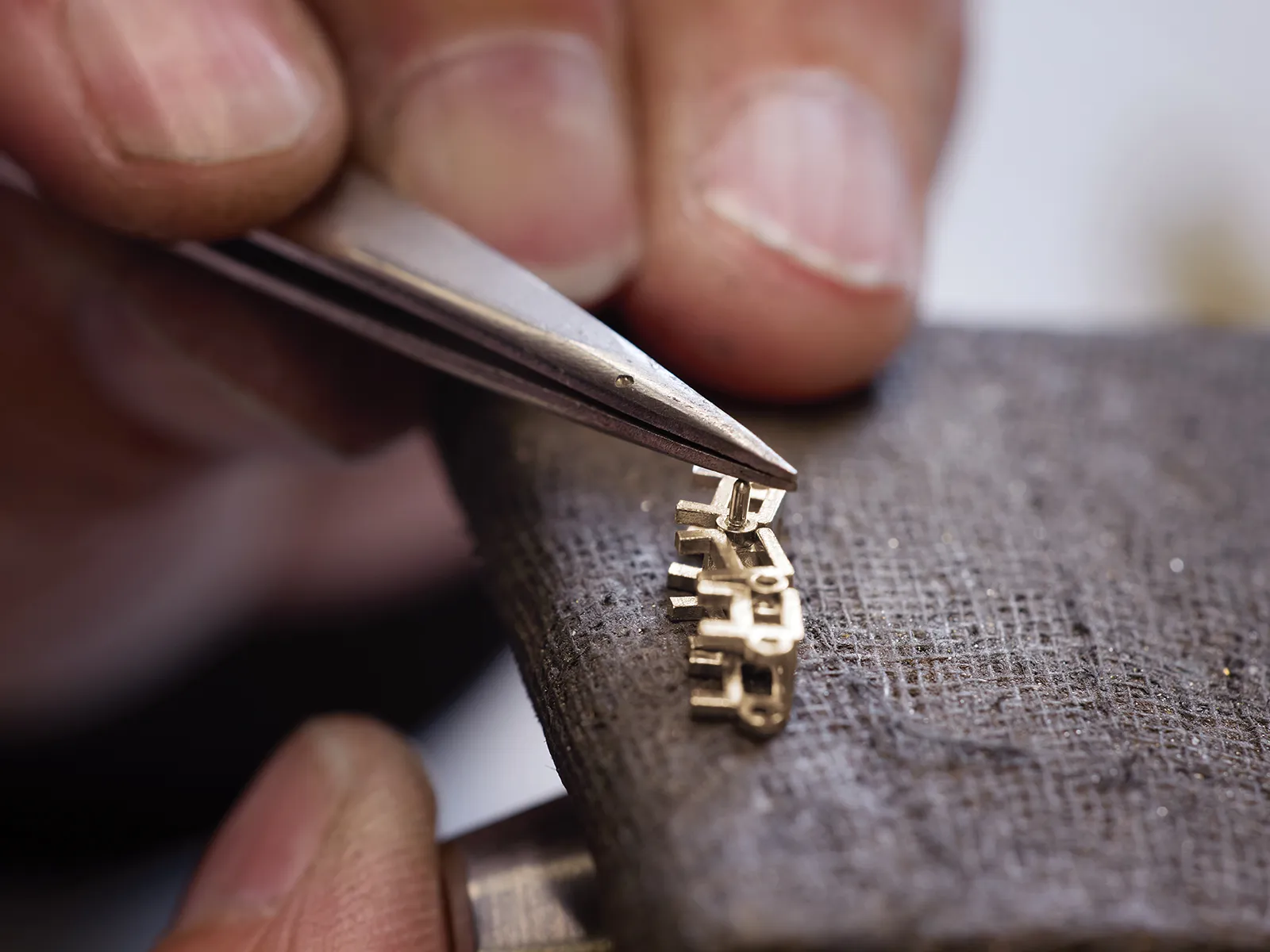 Close-up of hands using tweezers to work on small metal jewelry pieces on a textured surface.