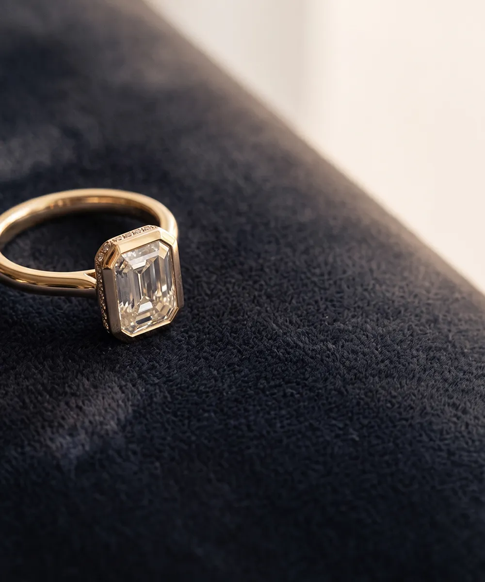 A gold ring with an emerald-cut diamond on a dark textured surface.