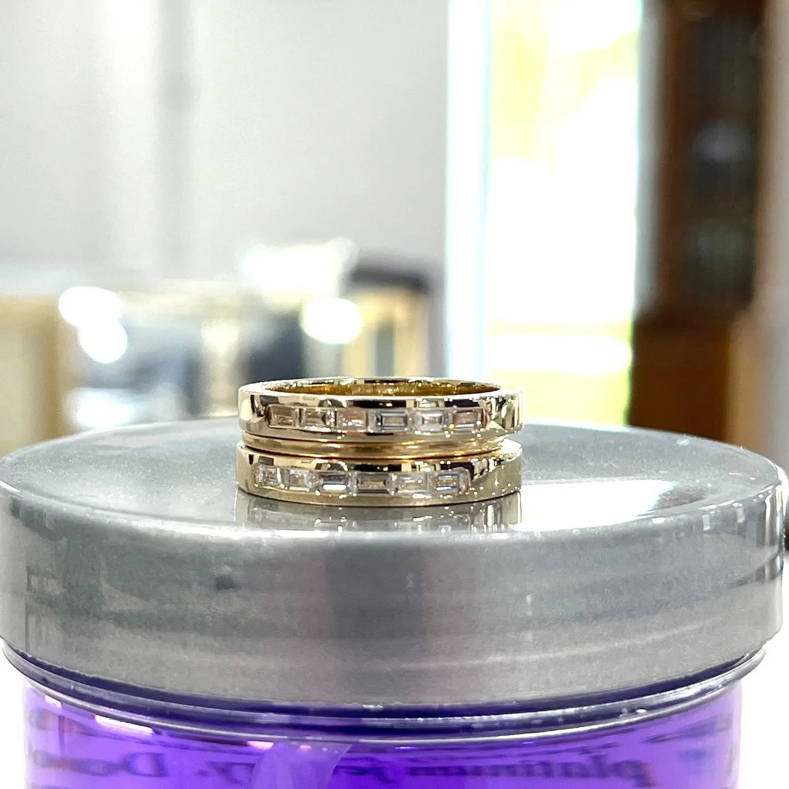 Two gold rings with rectangular diamonds on a reflective surface.