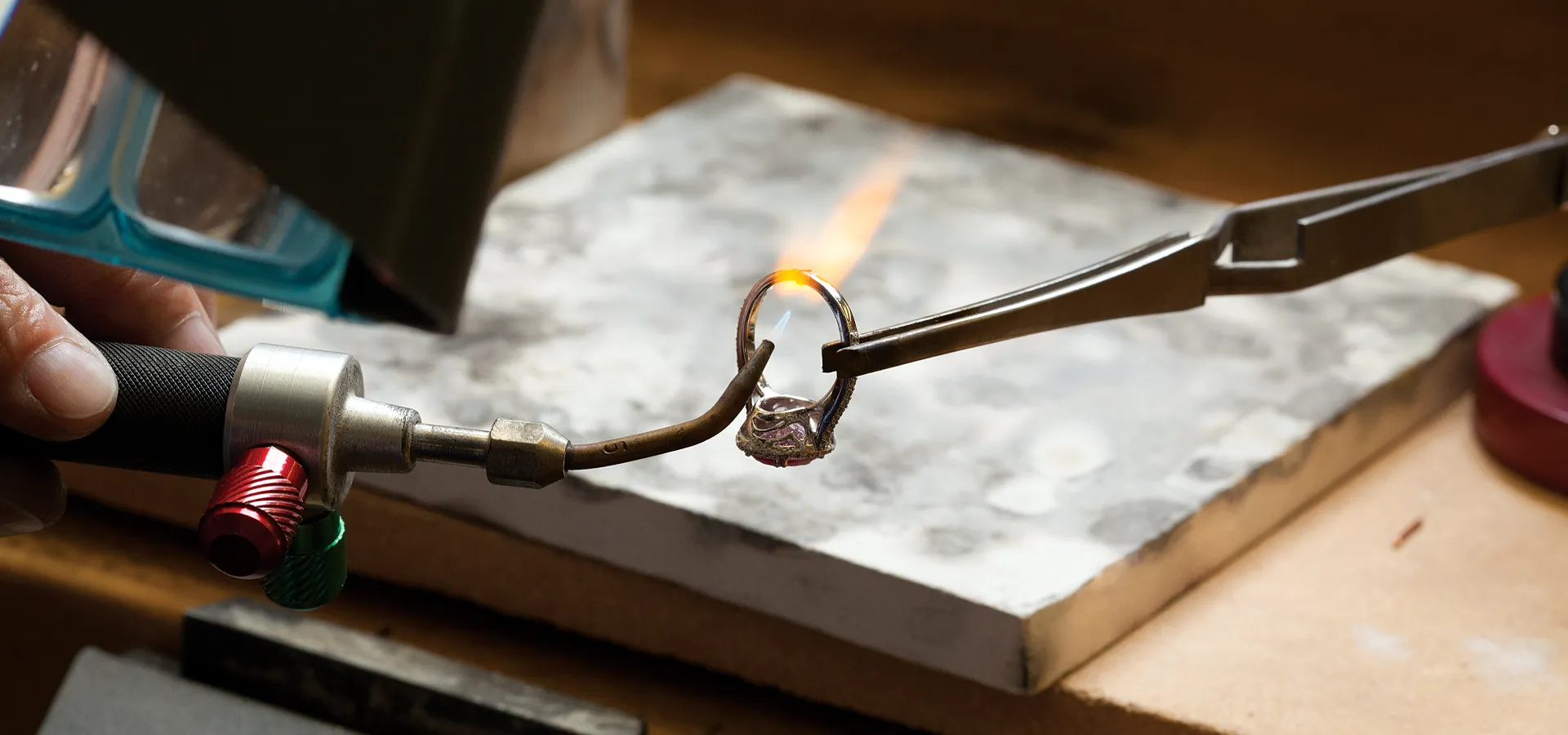 A jeweler uses a torch to solder a ring held by tweezers on a workbench.
