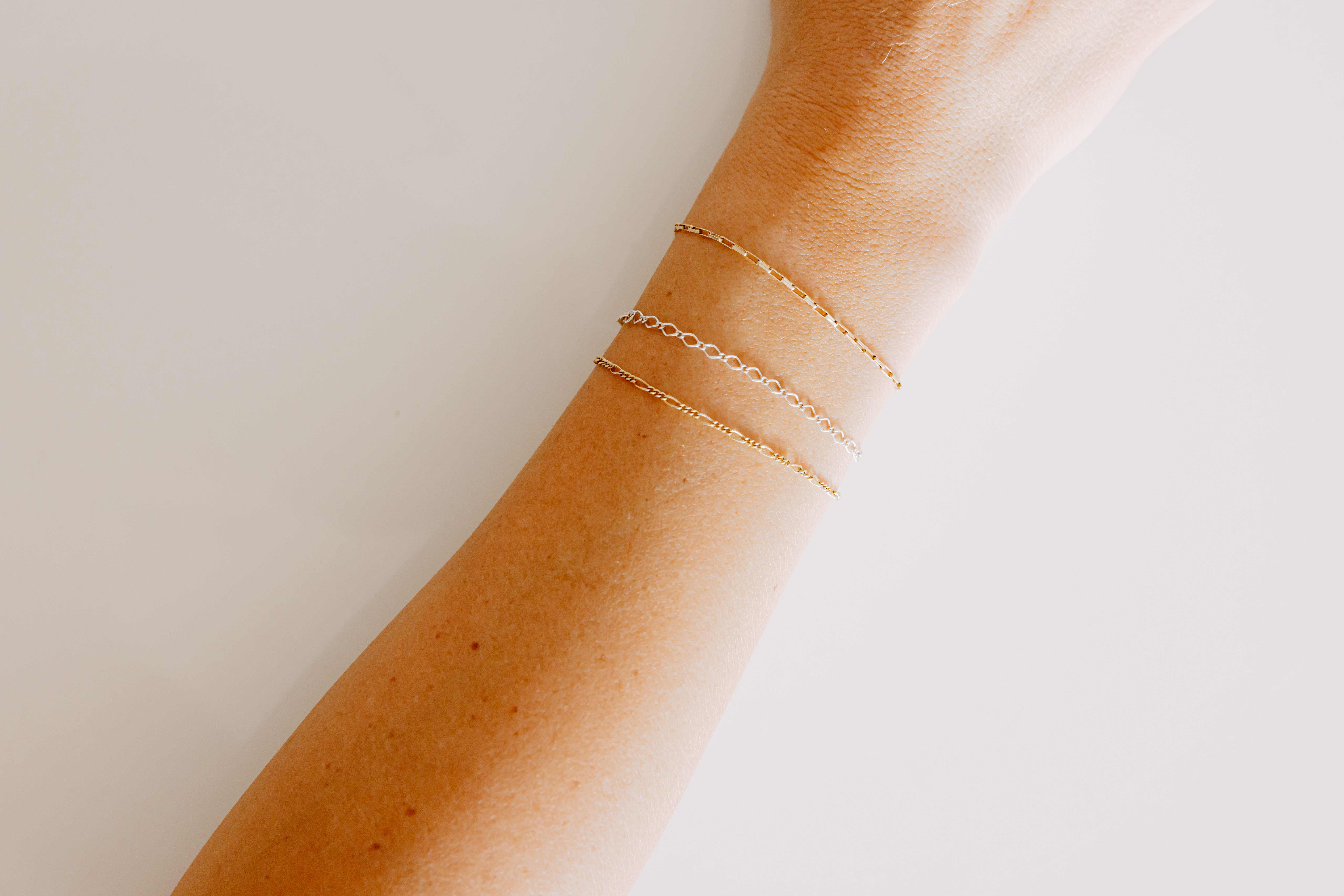 Three delicate permanent bracelets in gold and silver on a tanned arm against a light background. 