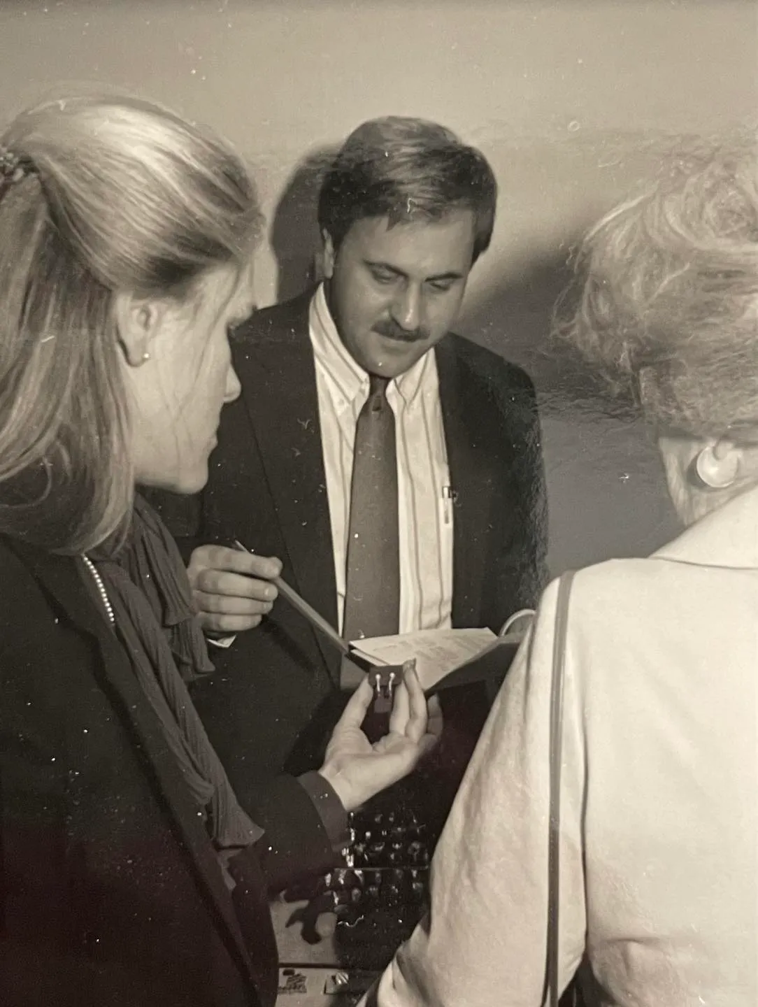 Three people discussing jewelry; one holds a small item while another takes notes.