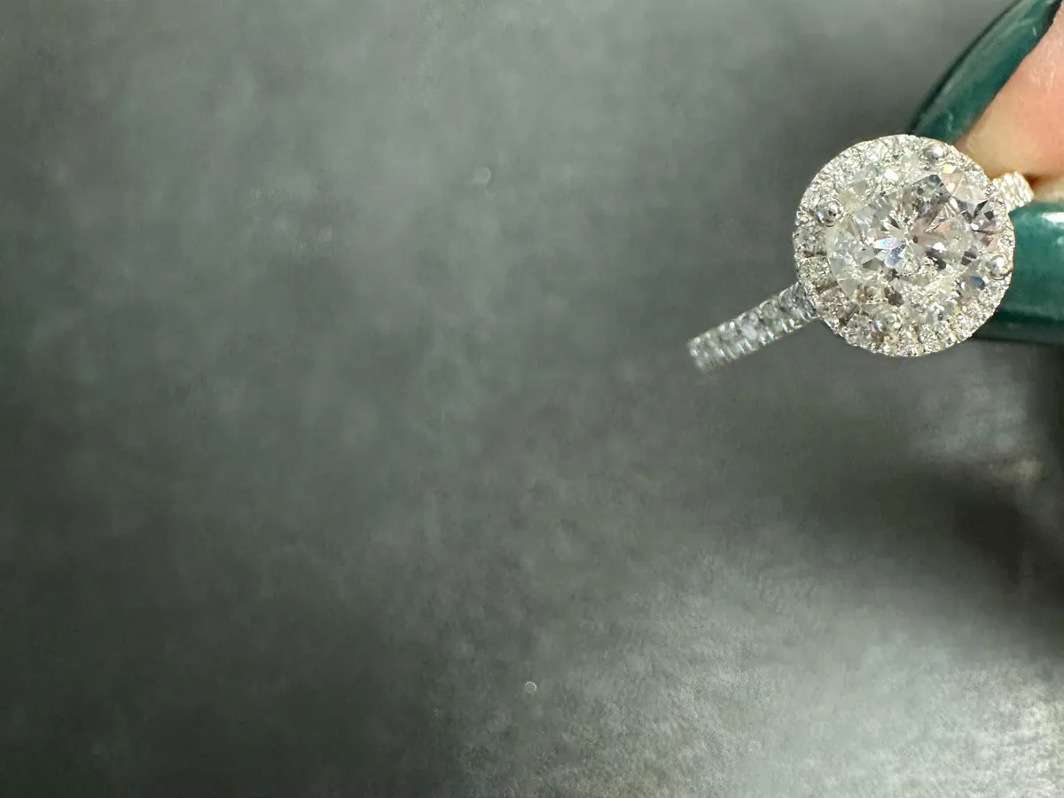 A sparkling diamond ring with a halo setting held by a hand against a dark background.