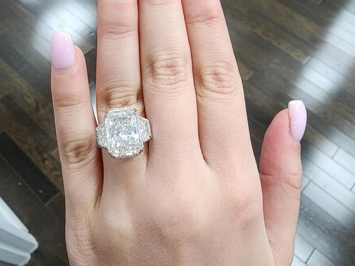 A hand with a large, radiant-cut diamond ring on the ring finger, nails painted light pink.