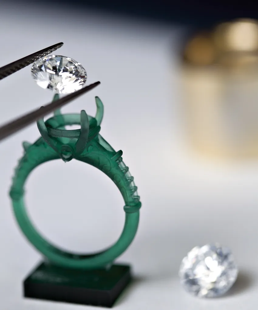 A diamond is held by tweezers above a green ring mold, with another diamond nearby.