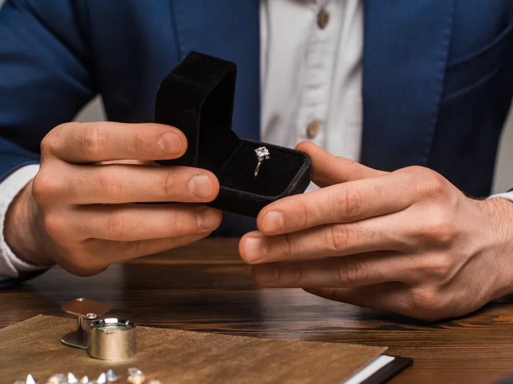 A person in a suit holds an open ring box with a diamond ring.