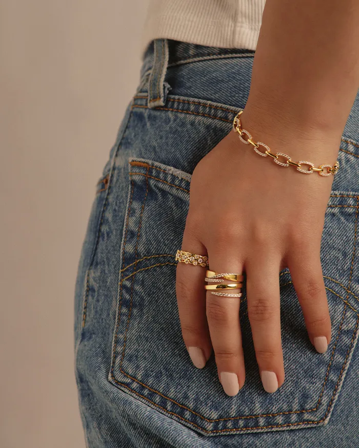 Close-up of a hand with gold rings and a bracelet, resting on denim jeans.