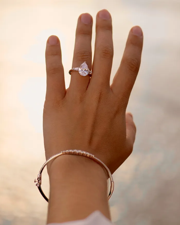 Hand displaying a pear-shaped engagement ring and a silver bracelet, with a soft background of water.