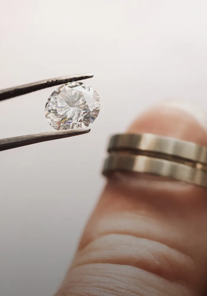 A hand holding tweezers with a diamond above a finger wearing two wedding bands.