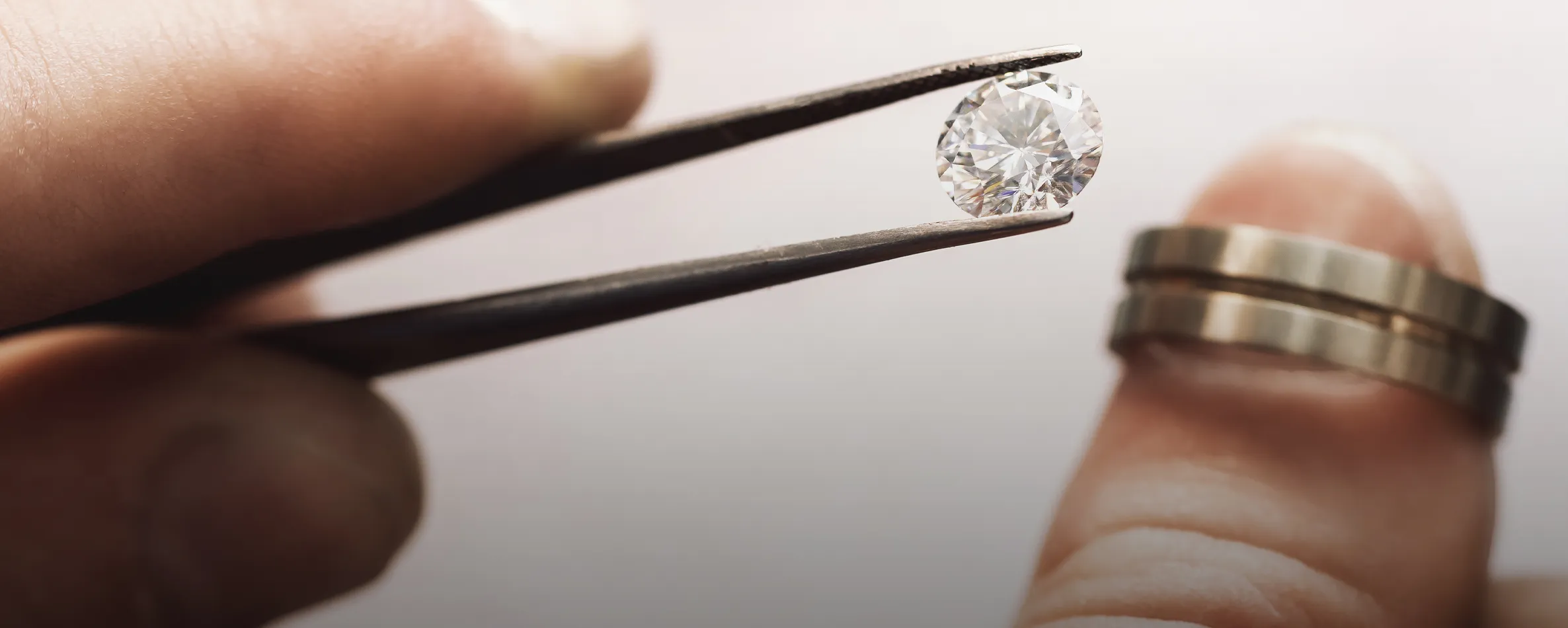 A hand holding tweezers with a diamond above a finger wearing two wedding bands.