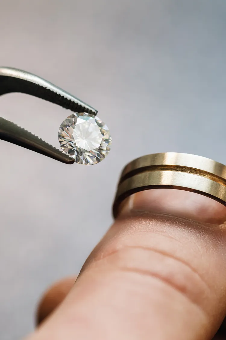 A hand holding a gold wedding band with a diamond being lifted by pliers, showcasing fine jewelry craftsmanship.