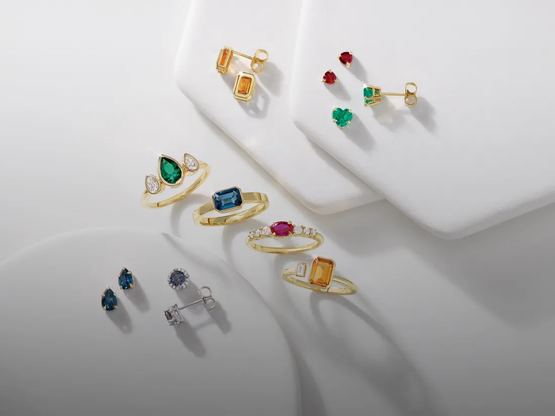 Colorful gemstone rings and earrings in gold settings, featuring various stones like emerald, ruby, and sapphire. 