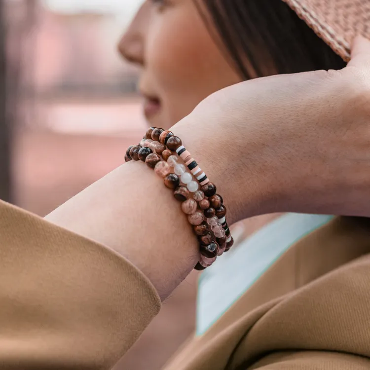 A person wearing stacked beaded bracelets, adjusting a hat outdoors. Click to Shop Now at Simones Jewelry.