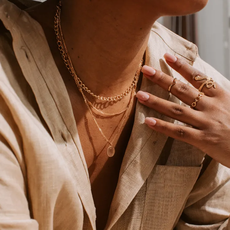 Layered gold necklaces and rings worn by a model in a light linen outfit. Click to shop at Simones Jewelry.