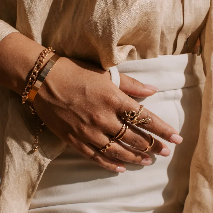 Layered gold rings and bracelets on a hand wearing a light linen outfit. Click to shop at Simones Jewelry.