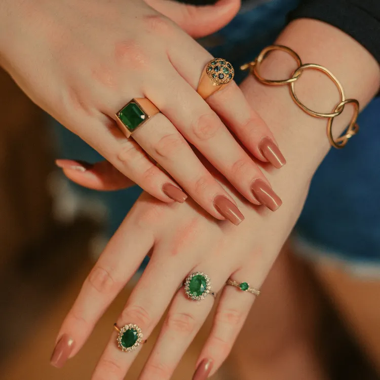 Hands adorned with green gemstone rings and a gold bracelet. Click to Shop Now at Simones Jewelry.