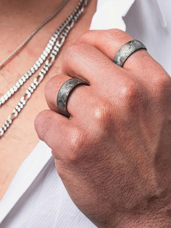 Close-up of a hand wearing two textured metal rings, with silver chains visible in the background. Shop fine jewelry at Simones 