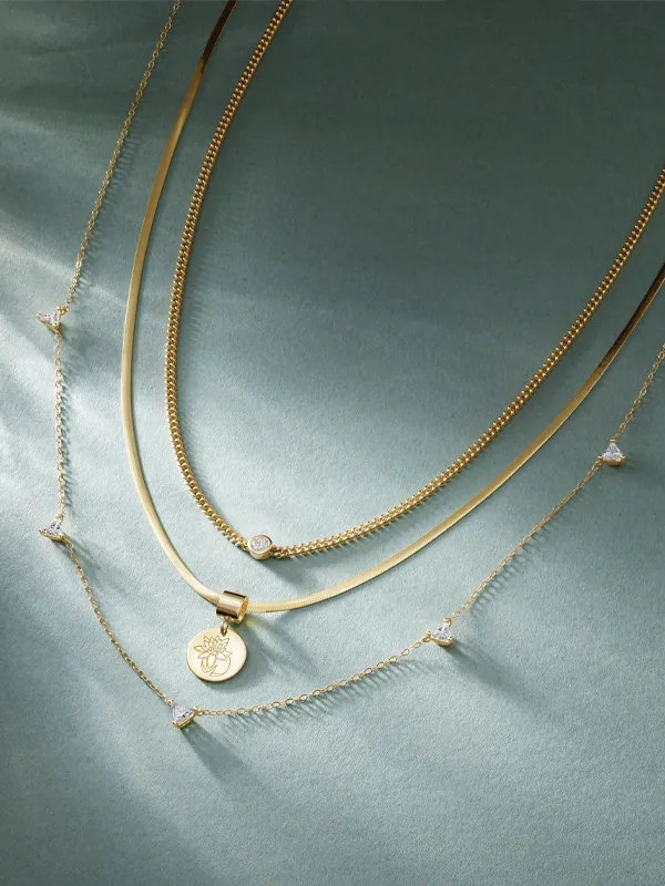 Three elegant gold necklaces with a pendant and diamond accents, displayed on a soft green background. Shop at Simones Jewelry.